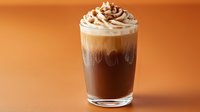 Layered iced coffee drink with whipped cream and chocolate sprinkles in clear glass against warm orange background, showcasing rich coffee colors and textures.
