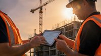 Two male construction workers in safety vests review building plans on a tablet at a job site with a crane and structure at sunset.