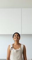 Young South Asian woman wearing a light linen apron over a white sleeveless top, smiling while looking upward in a bright, minimalist kitchen setting with white cabinetry.