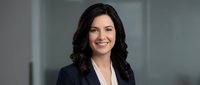 Professional businesswoman with dark wavy hair smiling confidently in navy blazer and white shirt, corporate headshot for marketing materials.