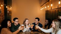 Group of diverse young adults toasting with beer glasses at indoor bar or restaurant with warm lighting and brick walls, celebrating together.