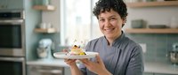 Chef with curly dark hair and warm smile holding plate with gourmet dessert in modern kitchen with green cabinetry and stainless steel appliances. Professional culinary presentation.