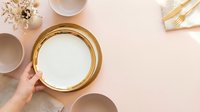 Woman's hand holding empty gold-rimmed plate on pastel pink background with dried flowers, cups and cutlery for table setting and event styling.