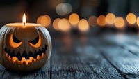 Glowing carved pumpkin with scary face on rustic wooden surface against blurred warm bokeh lights background creates spooky Halloween atmosphere.