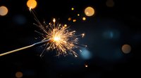 Glowing sparkler emitting bright golden sparks against dark background with bokeh lights, creating magical festive atmosphere for celebration and holiday designs.