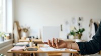 Woman with medium skin tone holding blank tablet device at wooden desk with natural light, office supplies, and plants for app mockup, web design, and digital product presentation.