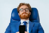 Young caucasian businessman with red beard and glasses relaxing in blue office chair while holding smartphone, wearing blue suit and tie against light background.