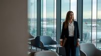 Professional businesswoman in dark suit standing in modern office with city view through floor-to-ceiling windows, corporate executive workspace.