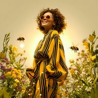 Young woman with curly brown hair and medium skin tone wearing yellow striped shirt and round sunglasses stands in blooming wildflower field with flying bees during golden hour.