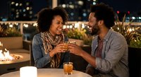 Happy African American couple toasting with drinks on romantic rooftop terrace at night with city lights and warm ambiance for dating lifestyle content.