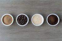 Four white bowls on wooden surface display coffee stages: cocoa powder, roasted beans, creamy espresso, and ground coffee in a row.