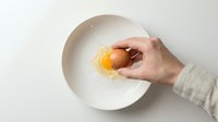Caucasian hand cracking raw egg into white bowl against neutral background, cooking preparation and food ingredient concept for culinary and kitchen-related projects.