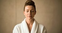 Middle-aged woman with fair skin and brown hair styled in an updo, wearing a white bathrobe, eyes closed in a serene expression against a neutral beige background for wellness and spa concepts.