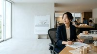 Young Asian female professional in dark blazer seated at wooden desk in modern office, reviewing architectural plans and building models, suitable for corporate, business, and design industry content.