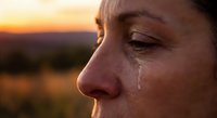 Close-up profile of woman with tear rolling down cheek during golden hour sunset, emotional moment capturing grief, sadness or contemplation in warm light.