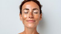 Young woman with freckles and closed eyes smiling peacefully against light background, showcasing natural beauty and skincare concept.