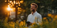 Smiling man with tablet enjoying sunset in meadow of yellow wildflowers, representing digital nomad lifestyle and work-life balance.