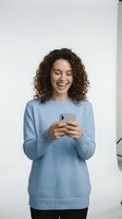 Young woman with curly brown hair wearing light blue sweater using smartphone, smiling while texting or browsing. Isolated on white background for app, website, and marketing use.