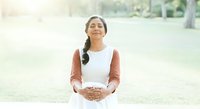 Young woman with dark braided hair and South Asian ethnicity wearing white sleeveless top with rust-colored cardigan, standing outdoors in park setting with hands clasped at stomach, smiling at camera.