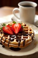 Fresh Belgian waffle topped with chocolate sauce and sliced strawberries served on white plate with coffee cup in background, morning breakfast setting.