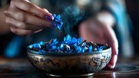 Mystical blue flowers in antique bronze bowl with rising smoke creating magical atmosphere. Woman's hand with purple manicure placing delicate petals.
