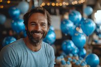 Mature bearded Caucasian man with genuine smile at celebration party with blue balloons and bokeh lights in background, wearing casual grey t-shirt.