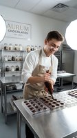 Male artisan chocolatier with light skin tone pouring melted chocolate into molds at professional confectionery workstation, wearing beige apron and white shirt in modern chocolate factory kitchen with ingredient shelves and equipment visible.