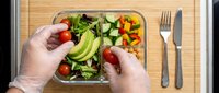 Hands preparing fresh salad with vegetables in glass container on wooden surface, healthy eating and meal prep concept.