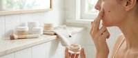 Young woman applying skincare cream in bright bathroom with natural light, holding jar of moisturizer for daily beauty routine and self-care ritual.