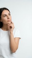 Thoughtful young woman in white t-shirt looking up with hand on chin in contemplative pose against neutral background for business and lifestyle concepts.