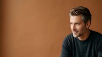 Middle-aged Caucasian man with dark hair and stubble wearing casual dark t-shirt against warm brown studio background, suitable for corporate portraits and professional headshots.