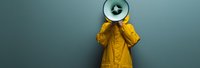 Person wearing yellow raincoat holding megaphone against gray wall background, concept of public announcement, protest, or communication in minimal style.