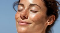 Young Caucasian woman with freckles and glowing skin enjoying sunlight with eyes closed, expressing serenity and natural beauty against blue sky background.