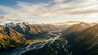 Aerial view of winding river flowing through alpine valley surrounded by snow-capped mountains during golden hour sunset, showcasing dramatic landscape and natural terrain for travel and nature projects.