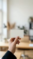 Hand holding blank white business card in modern office workspace with natural light and blurred background, suitable for branding and corporate mockups.