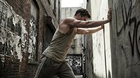 Muscular young Caucasian man leaning against graffiti wall in urban alley, wearing tank top, dramatic street atmosphere, gritty city background.