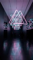 Modern neon geometric triangular light installation hanging in dark contemporary gallery space with purple and cyan illumination creating futuristic atmosphere.