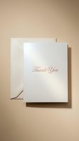 Elegant white thank you card with rose gold script lettering and beige envelope on warm neutral background, for weddings and events.