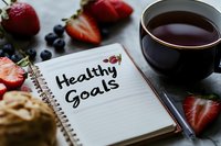 Notebook with healthy goals text surrounded by fresh strawberries, blueberries and cup of tea, wellness and diet planning concept on wooden table.