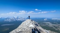 Lone hiker stands triumphantly on rocky mountain peak overlooking vast mountain range and valleys below, dramatic landscape under blue sky with scattered clouds.