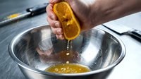 Male hand squeezing fresh orange half over stainless steel bowl with juice dripping, citrus preparation for cooking or beverage making on kitchen counter.