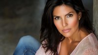 Confident Hispanic woman with long dark hair wearing pink sweater and jeans, sitting casually against blurred background in natural lighting for lifestyle portrait.