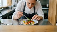 Chef plating gourmet dish with fresh vegetables and garnish in professional kitchen. Fine dining food preparation and culinary artistry.