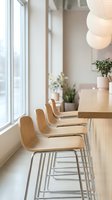 Modern wooden bar stools with metal frames lined along a light wood counter near bright windows in a minimalist kitchen or cafe space with pendant lighting and potted plants.