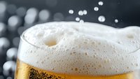 Macro shot of fresh beer with white foam head and rising bubbles against dark background, showing carbonation and refreshing quality of cold draft lager beverage.