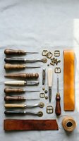 Leather crafting tools flat lay with wooden handles, awls, stitching wheels, buckles, thread, and tan leather strip on linen background.