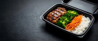 Grilled meat with broccoli, carrots and rice in black compartmented takeout container on dark surface for food delivery and meal prep.