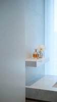 Amber perfume bottle with golden cap displayed on white floating shelf beside ceramic diffuser with white petals in minimalist bathroom setting with natural light.