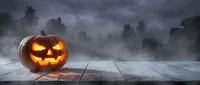 Glowing jack-o-lantern pumpkin with carved scary face on wooden table against spooky foggy cemetery background for Halloween celebration and autumn decor.
