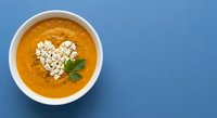 Creamy orange soup with heart-shaped croutons and fresh herbs in white bowl on blue background for romantic dining and comfort food concepts.